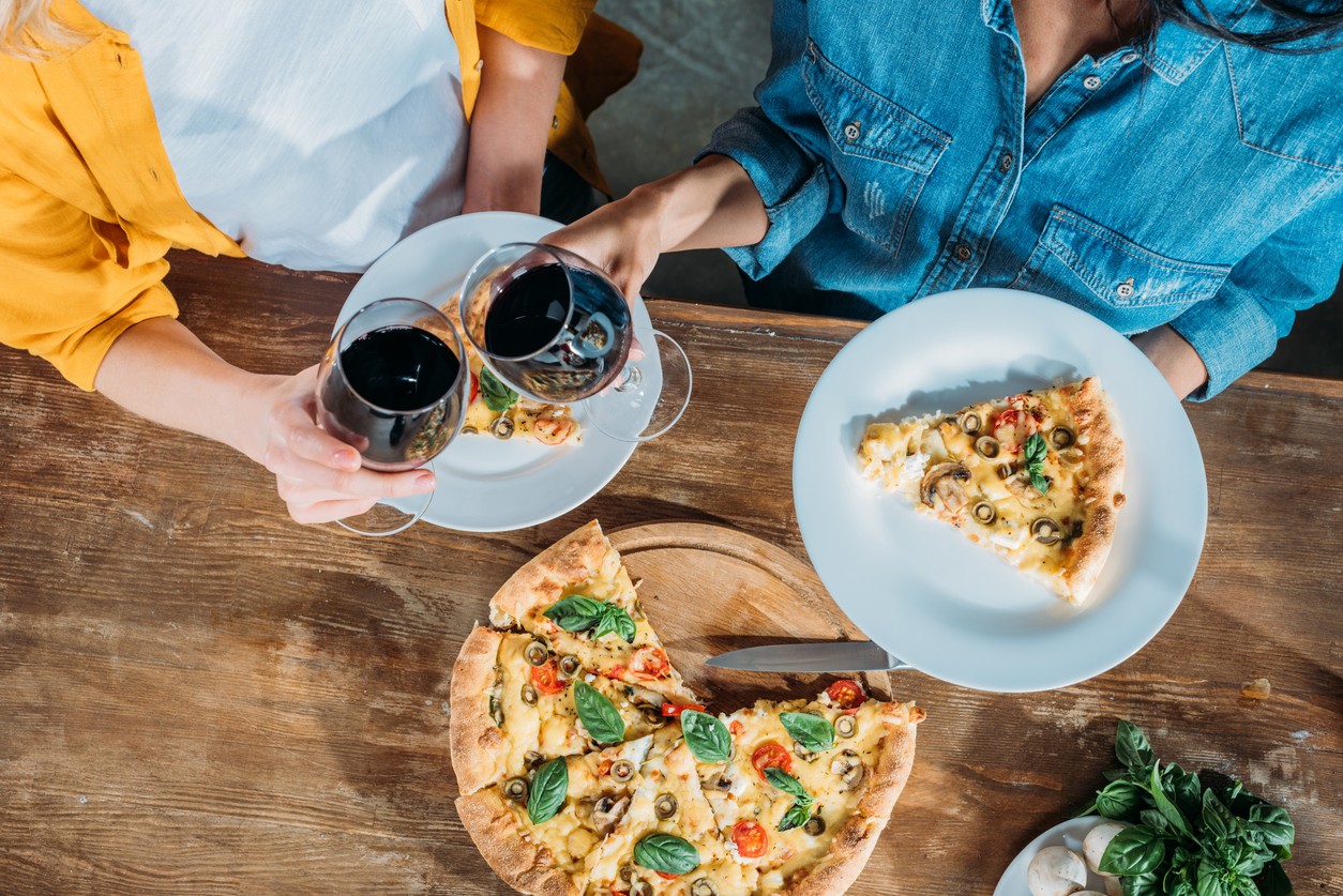 Top view of young women clinking wine glasses while eating homemade pizza together (Foto: Getty Images/iStockphoto)