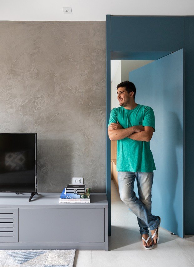 O morador de 23 anos posa em frente ao painel de marcenaria azul que camufla a porta de seu quarto (Foto: Lufe Gomes/Divulgação)
