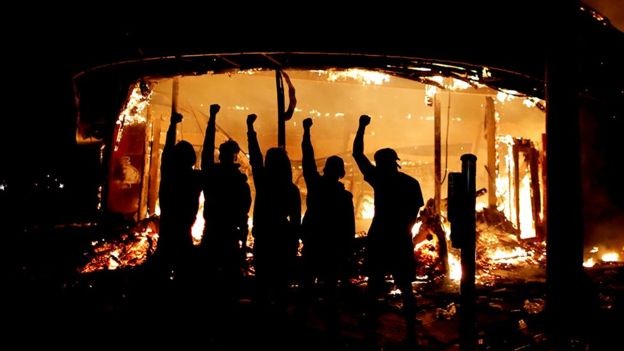 Manifestantes posam do lado de fora de loja de bebidas em chamas  (Foto: Reuters via BBC)