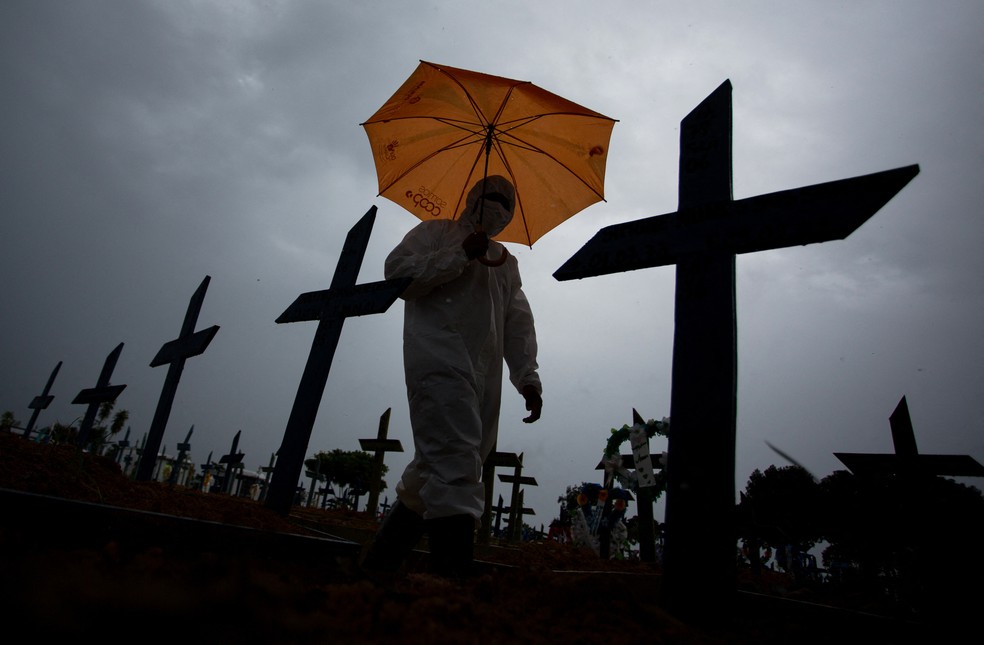 Funcionário anda com roupas protetoras e um guarda-chuva em meio a túmulos no cemitério Nossa Senhora Aparecida, em Manaus, no dia 25 de fevereiro. — Foto: Michael Dantas/AFP