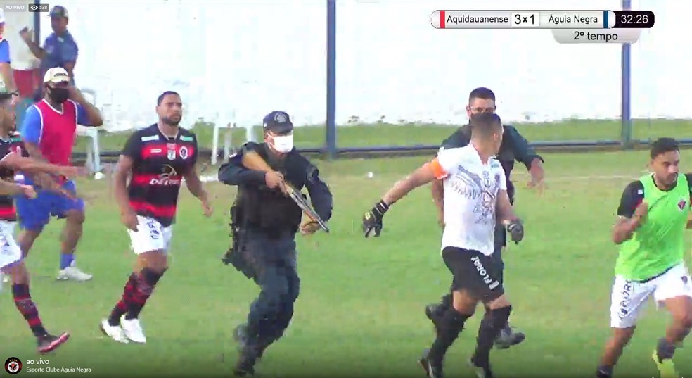 Momento em que policial, com arma na mão, persegue o zagueiro Virgulino, no estádio Noroeste, em Aquidauana — Foto: TV Águia/Reprodução