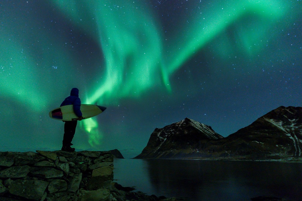 Surfista aprecia as luzes do norte em Utakleiv, norte da Noruega, no arquipélago de Lofoten  (Foto: Olivier Morin/AFP)