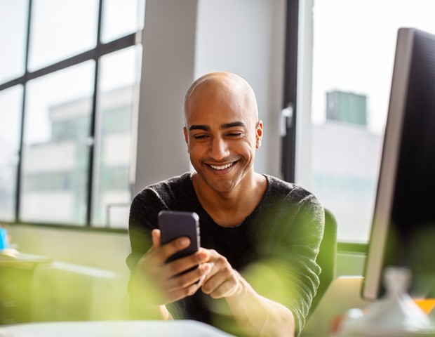 Smiling mid adult man using phone and smiling. Hispanic male professional texting on his cell phone while working at his desk. (Foto: Getty Images)
