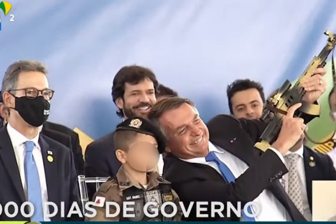 Bolsonaro fez poses segurando o fuzil de brinquedo (Foto: Reprodução Tv Brasil)