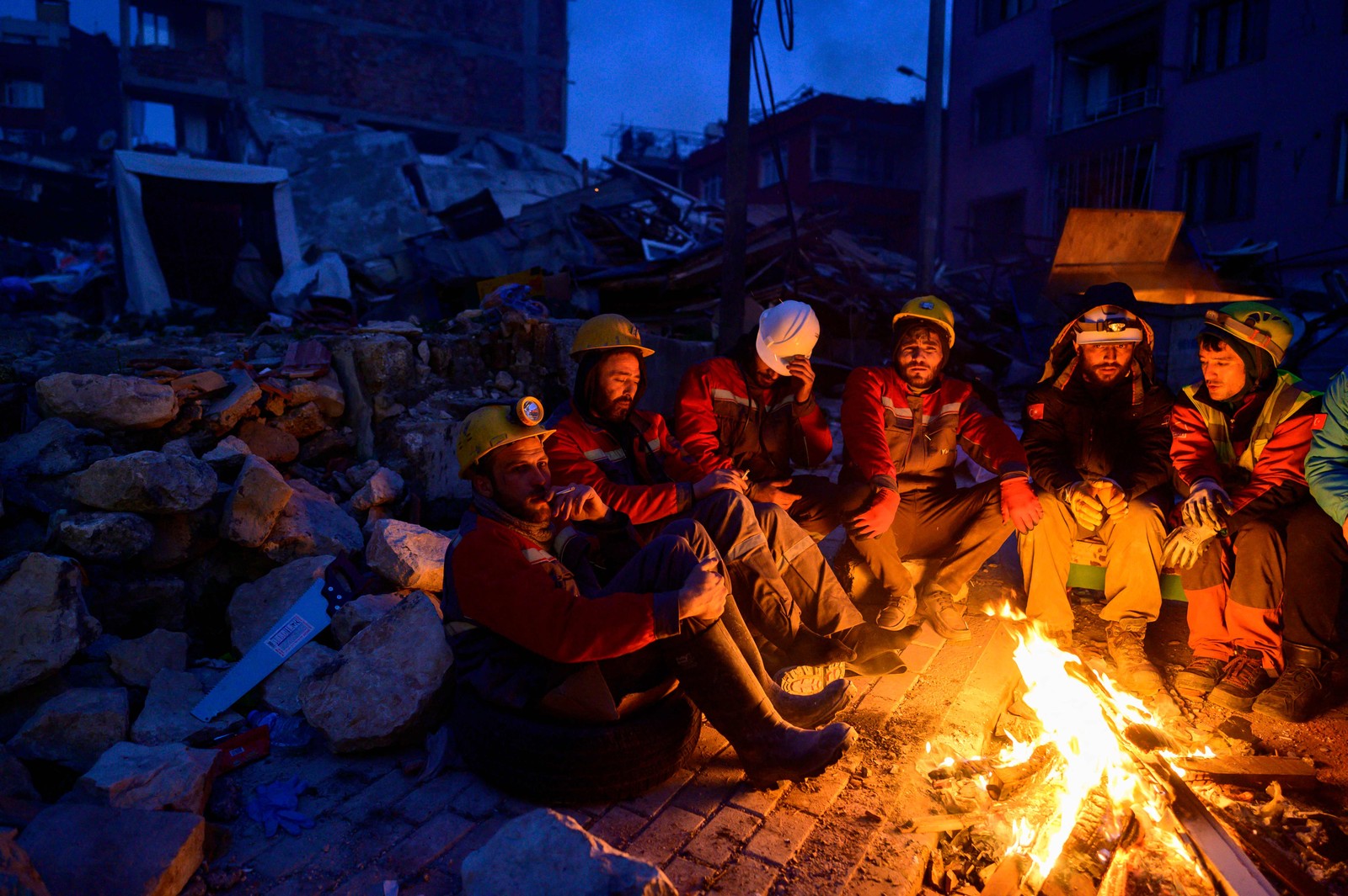 Membros de equipes de resgate se sentam em torno de fogueira para se aquecer sobre os escombros — Foto: YASIN AKGUL/AFP