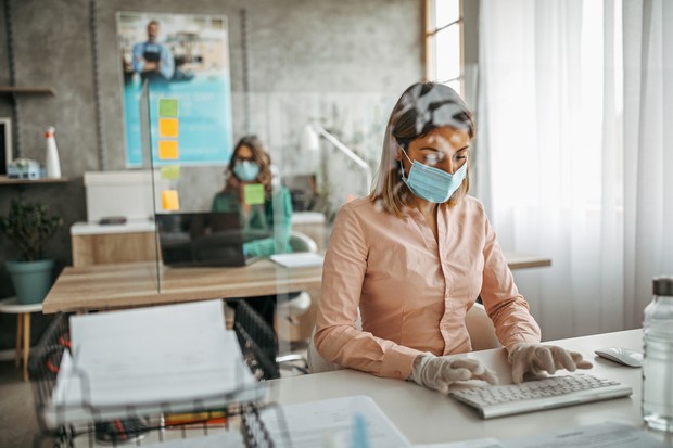 Two businesswomen working at office wearing protective gloves and face mask maintaining social distance. Office with acrylic glass partition on desk. Acrylic glass wall - protection against coughs and spitting, protection against viruses. (Foto: Getty Images)