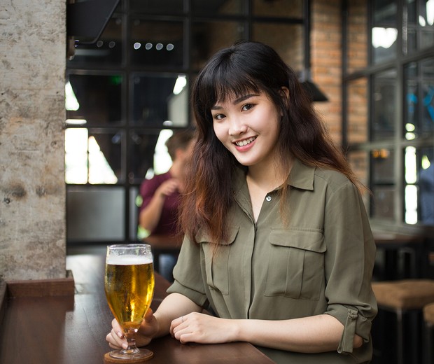 Closeup portrait of smiling young Asian woman looking at camera and sitting at table with glass of beer in pub with blurred customers in background (Foto: Divulgao/Freepik)