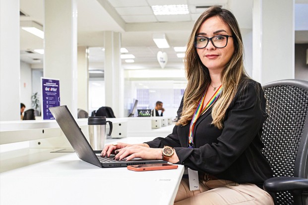 Foto de Diana Fonseca Resende sentada em um escritório com as mãos sobre o teclado de um laptop. Ela é branca, tem cabelos lisos e longos e está com um óculos de grau. 