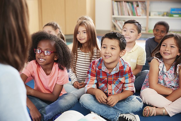 Elementary school kids sit on floor looking up at teacher (Foto: Getty Images/iStockphoto)