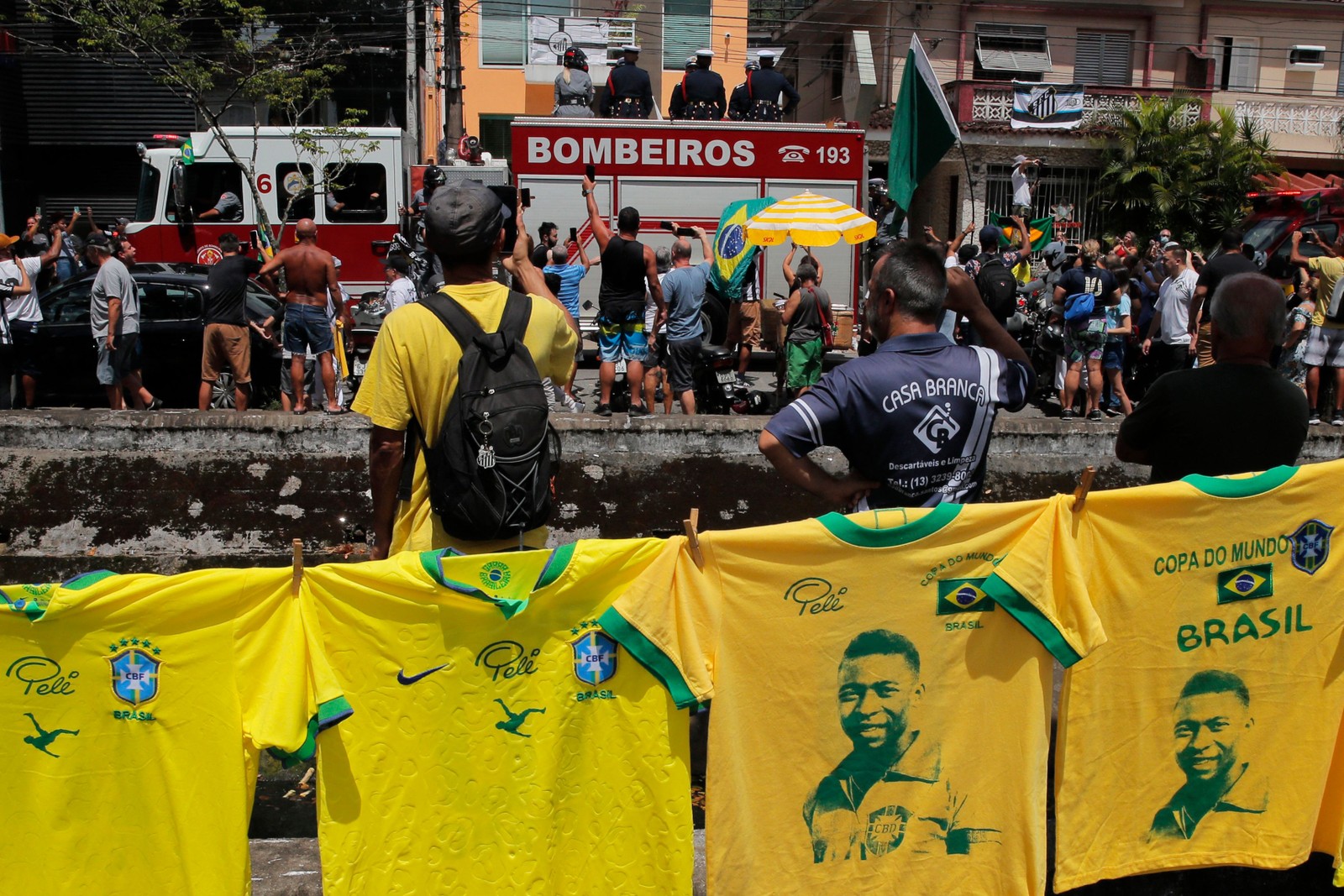 Camisetas lembrando o falecido astro do futebol brasileiro penduradas em uma rua enquanto um caminhão de bombeiros transporta o caixão de Pelé para o Cemitério Memorial de Santos — Foto: CAIO GUATELLI / AFP