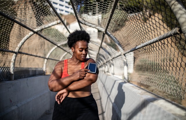 Healthy woman touching phone screen on armband before exercising outdoors. African american woman in sports clothing using phone while exercising outdoors. (Foto: Getty Images)