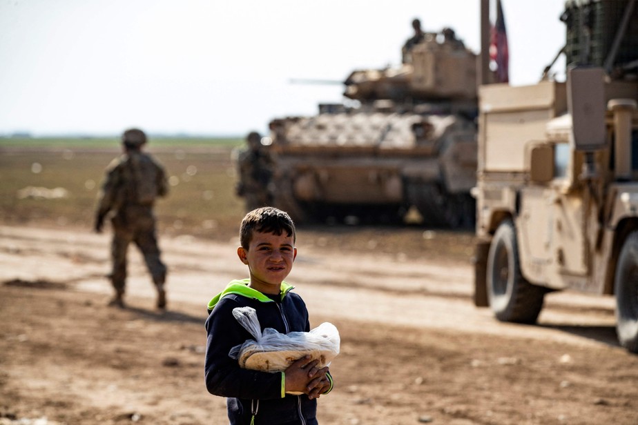 Criança carregando uma sacola com pão ao lado de tropas americanas na cidade de Tal Hamis, na Síria