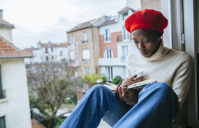 Young african american woman sitting in a window with a book and a cup. In Paris, France. (Foto: Getty Images)