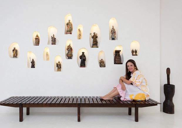 No hall de entrada, parede com coleção de santos de madeira e marfim dos séculos 18 e 19, Malu sentada no banco de Sergio Rodrigues, usa blusa e saia, ambas Isabela Capeto (Foto: Xico Diniz)