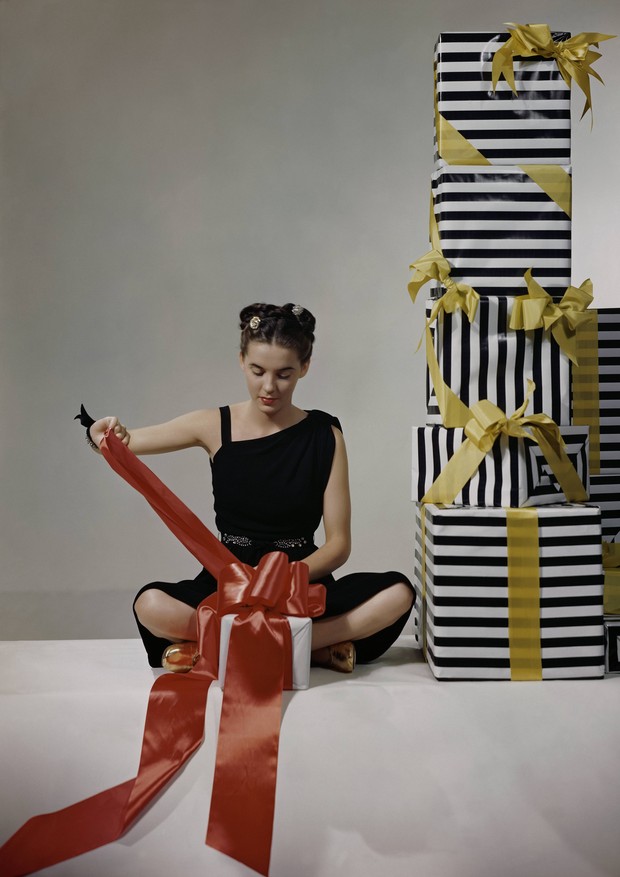 Model in black rayon dress by Margot with toga style shoulder, and sparkling belt, wrapping a gift next to pile of boxes. (Photo by Clifford Coffin/Condé Nast via Getty Images) (Foto: Conde Nast via Getty Images)