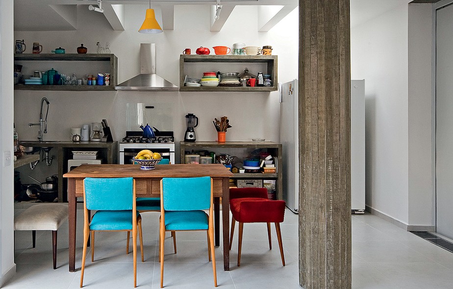 As cadeiras com tecido azul dão cor à cozinha neutra. Na cabeceira, há apenas um banco, no mesmo material, mas vermelho. Projeto do escritório Casa 14 Arquitetura