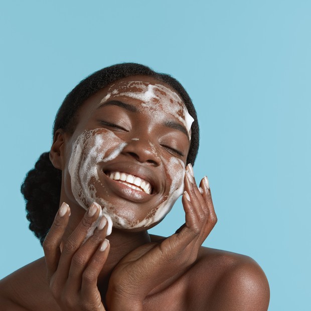 Close up portrait of beautiful black girl wash her face with cleansing face foam. Smiling young woman with closed eyes. Concept of face skin care. Isolated on blue background. Studio shoot (Foto: Getty Images/iStockphoto)