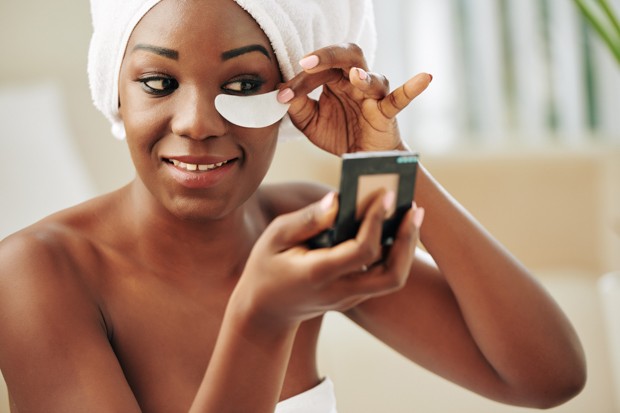 Horizontal close up portrait of beautiful young Black woman looking in pocket mirror applying eye patch (Foto: Envato)