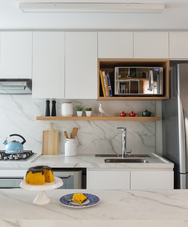 A cozinha recebeu revestimentos bem clean: porcelanato branco marmorizado cobre o piso, bancadas e frontão. O resto dos móveis da cozinha é branco, com detalhes em madeira que conversam com o apartamento (Foto: Manu Oristanio/Divulgação)