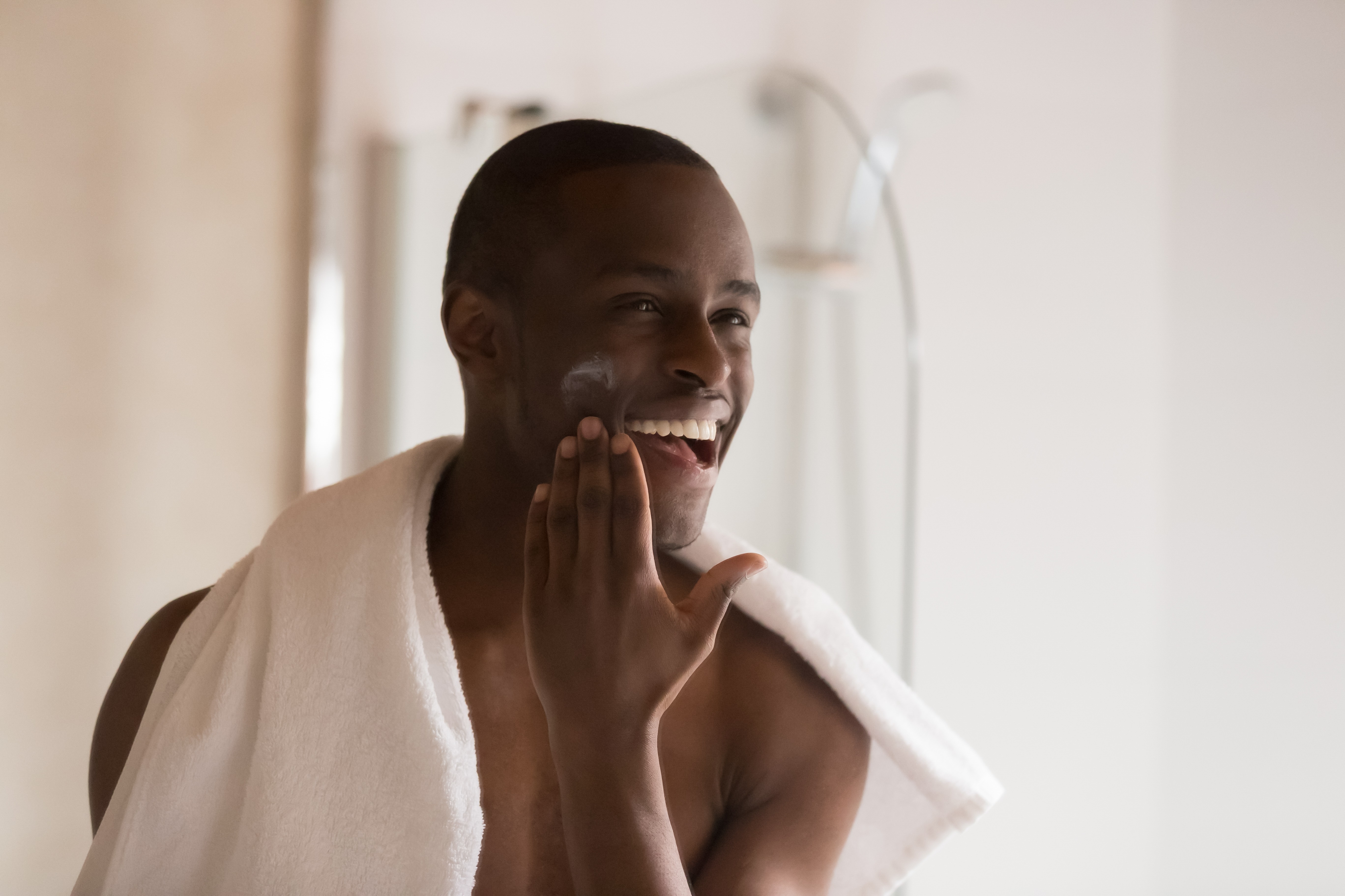 Happy smiling african ethnicity man looking in mirror at bathroom after showering, applying moisturizing after shaving balm on cheek, soothing and soften sensitive skin. Morning hygiene routine. (Foto: Getty Images/iStockphoto)