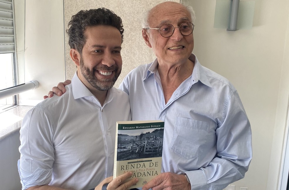 André Janones (à esq.) segura livro sobre Renda de Básica de Cidadania com Eduardo Suplicy — Foto: Reprodução