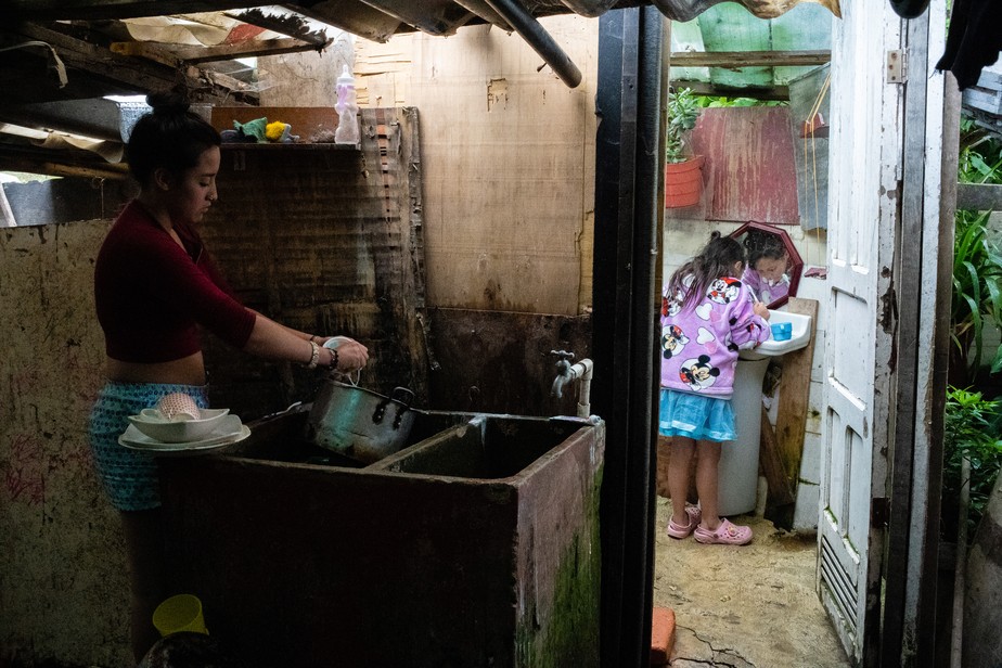 Garota lava pratos enquanto sua irmã mais nova escova os dentes em sua casa em Soacha, Colômbia