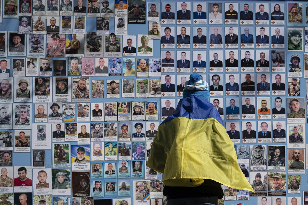 Mulher com a bandeira ucraniana nas costas olha para mural com as vítimas em Kiev, na Ucrânia — Foto: Andrew Kravchenko/AP