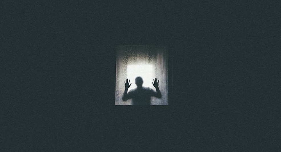 Imagem quase toda preta mostra, ao centro, silhueta de pessoa com as duas mãos apoiadas em um vidro. — Foto: Pexels