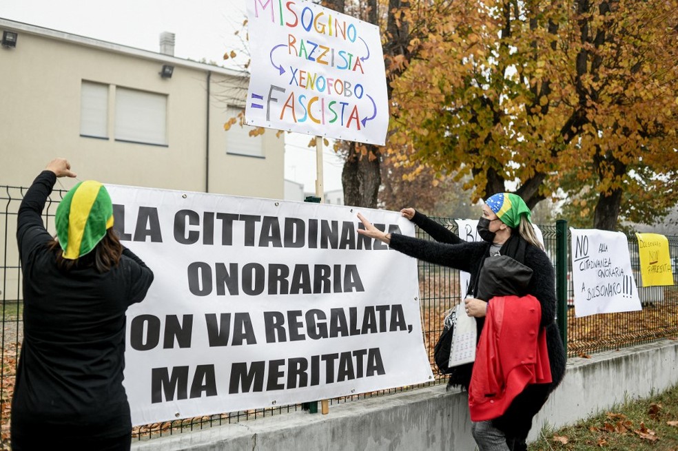Em  Anguillara Veneta, mulheres fixam faixa em italiano onde se afirma que o título de cidadão honorário não deveria ser dado como um presente, mas, sim, conquistado; manifestantes protestaram contra o presidente Jair Bolsonaro, em 1º de novembro de 2021 — Foto: Piero Cruciatti/ AFP