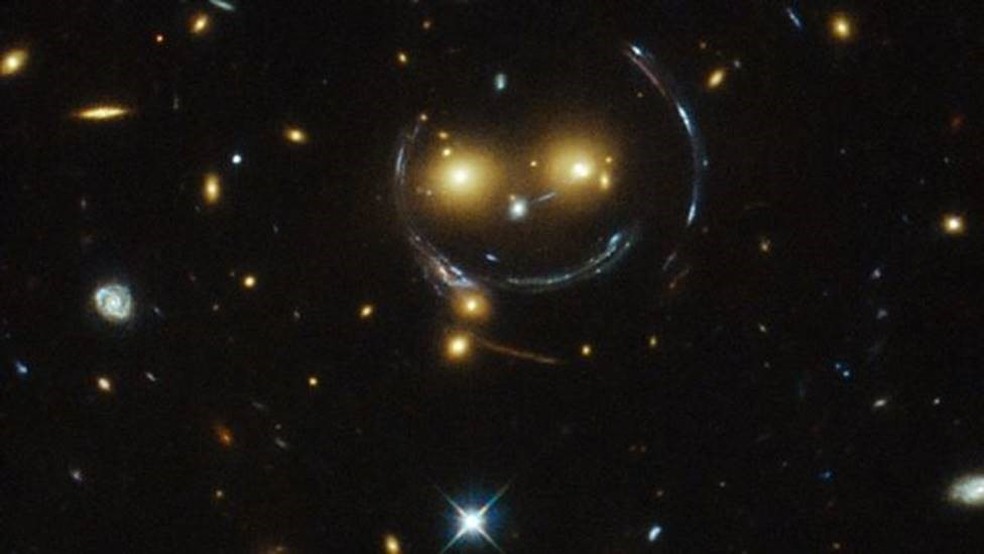 O aglomerado SDSS J1038+4849, em imagem captada pelo Hubble. No que parece um rosto sorridente no centro da imagem, os dois 'olhos' são galáxias muito brilhantes e o 'sorriso' é um arco de luz causado pelo efeito de lente gravitacional. O aglomerado distorce e curva a luz dos objetos que estão atrás dele — Foto: NASA