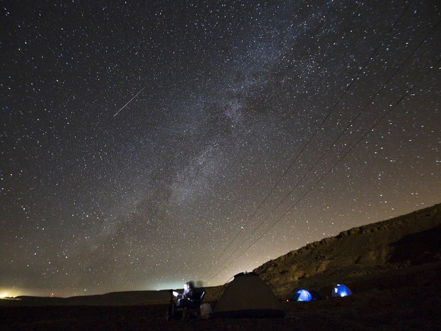 Pessoas acampam em lugar aberto à espera da chuva de meteoros Perseidas enquanto uma estrela cadente cruza o céu sobre Ramon Carter, em Israel. A mancha da Via Láctea também é vista no céu (Foto: Amir Cohen/Reuters)