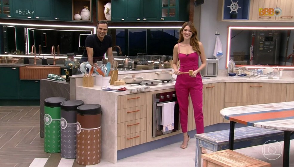 Na cozinha do BBB23 estão Tadeu e Ana Clara. Em destaque, a frente na foto, estão três lixeiras: uma verde, uma cinza e uma marrom.