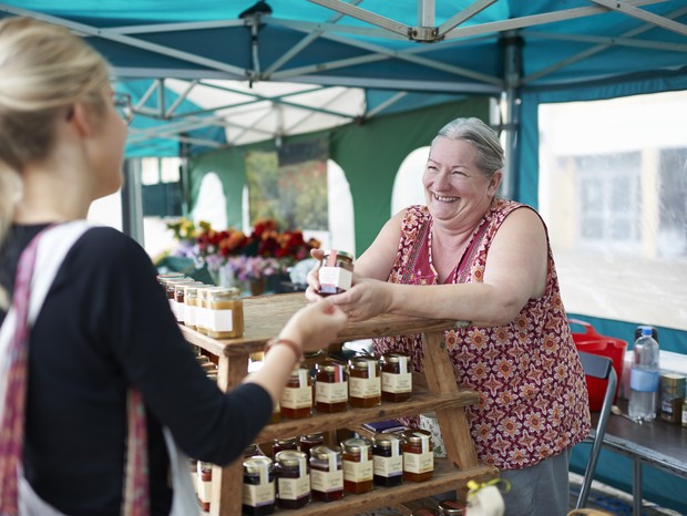 A senior woman working on a local market stall selling homemade jam serves a customer. (Foto: Getty Images)