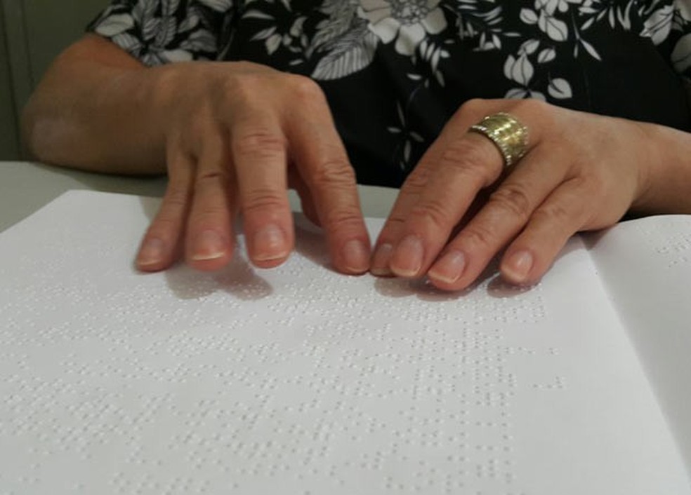Mulher usando as duas mÃ£os para ler uma pÃ¡gina de papel escrita usando o sistema braille: ao contrÃ¡rio de tinta, a mÃ¡quina de escrever em braille perfura o papel para criar os relevos respectivos de cada letra, nÃºmero ou pontuaÃ§Ã£o â Foto: Raquel Morais/G1