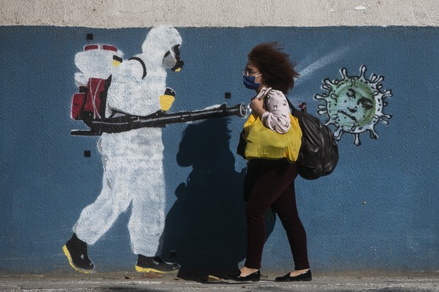 Uma mulher usando máscara passa por uma parede com um grafite que retrata um homem com equipamento de proteção espalhando vírus com o rosto do presidente Jair Bolsonaro no bairro Estácio, Rio de Janeiro, em junho de 2020 (Foto: Bruna Prado/Getty Image)