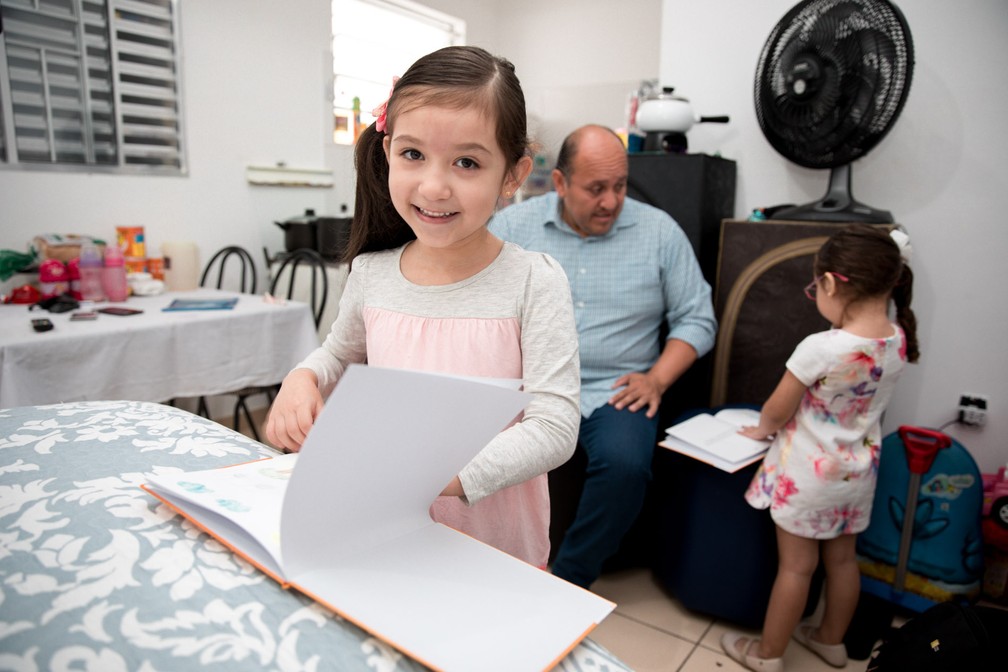 Valentina e sua irmã mais nova, Rafaela (ao fundo), folheam exemplares do livro escrito por ela na casa da família, no Brás, sob o olhar do pai, Cristiano (Foto: Marcelo Brandt/G1)