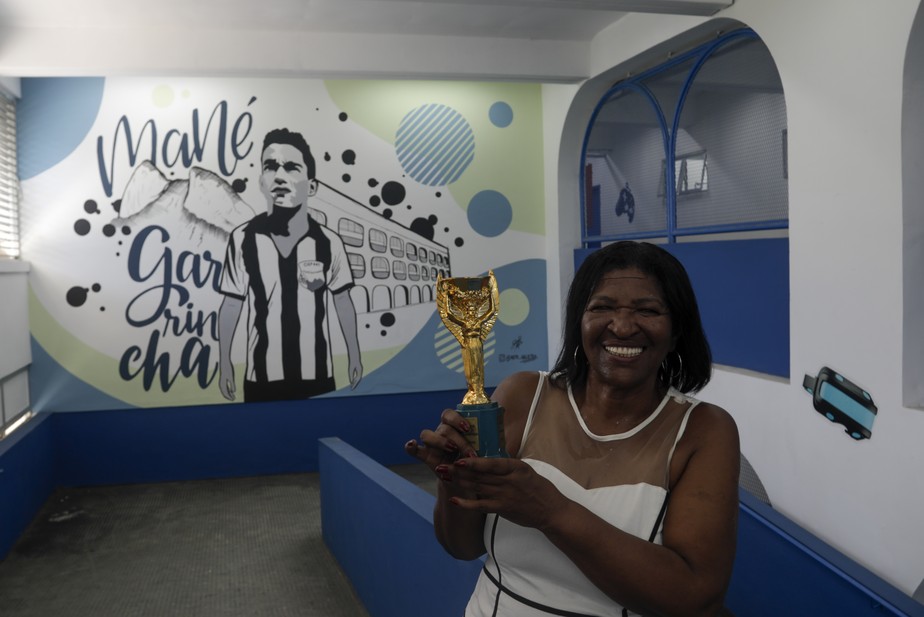 Filha de Garrincha, Rosângela dos Santos posa em frente ao grafite do pai em escola de Pau Grande; cidade possui tour que passa por locais ligados ao ídolo