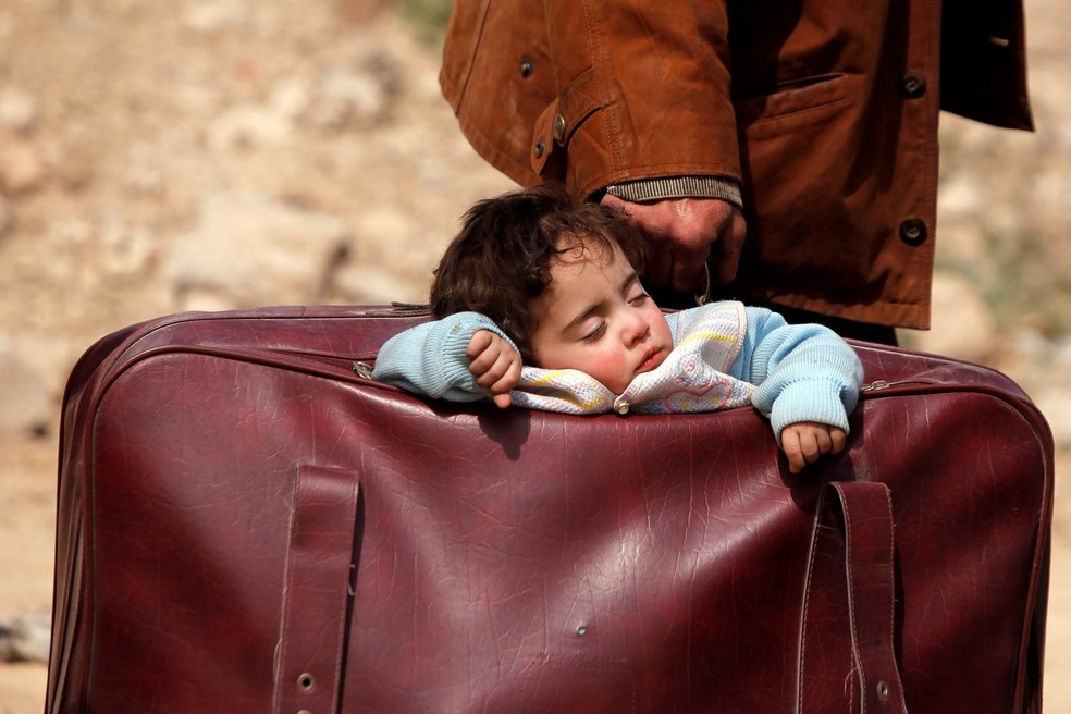 Homem leva um bebê dormindo dentro de uma mala de roupas em meio a dezenas de pessoas que abandonam a vila de Beit Sawa, no leste de Guta, na Síria, devido à guerra que assola a região (Foto: Omar Sanadiki/Reuters)