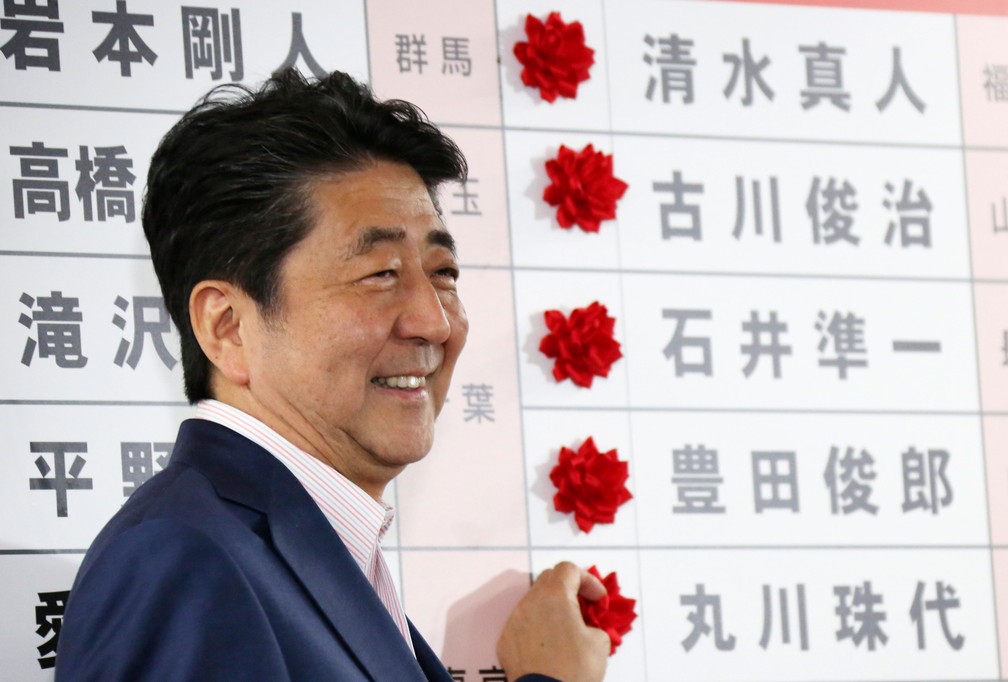 Primeiro-ministro japonês, Shinzo Abe, sorri neste domingo (21) diante das rosas vermelhas colocadas diante dos nomes dos candidatos vencedores do seu partido, o Liberal Democrata, das eleições para a Câmara Alta em Tóquio   — Foto: Koji Sasahara/AP
