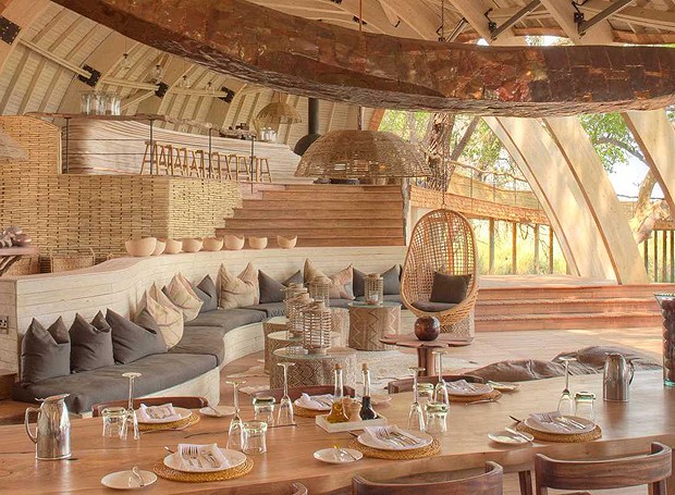 Área social na parte interna do hotel, com mesa de jantar, sofá e bar. Praticamente tudo foi feito com madeira no Sandibe Okavango Safari Lodge e isso o tornou um local mais aconchegante e confortável! (Foto: Divulgação)