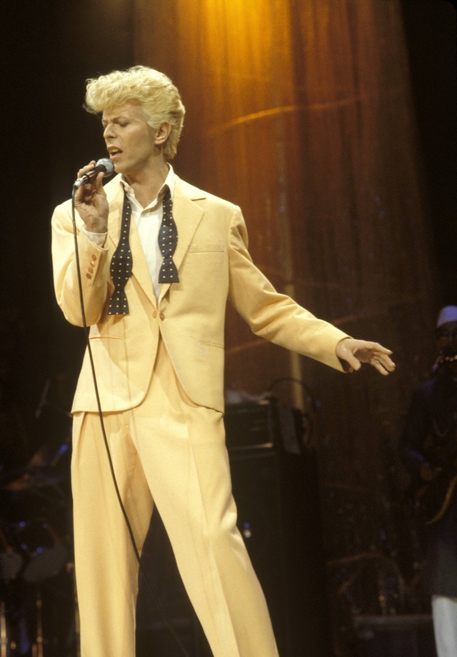 David Bowie de terno amarelo, cantando com microfone na mão na frente de uma cortina com luz bege (Foto: Getty Images)