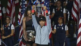 Naomi Osaka levanta a taça de campeã do Aberto dos EUA após vencer disputa contra Serena Williams (ao fundo) — Foto: Jean Catuffe/Getty Images