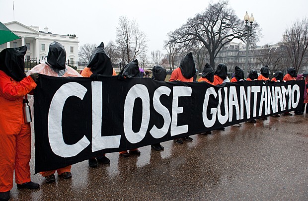 Manifestantes com macacões alaranjados de presidiários e capuzes pretos seguram faixa que pede ‘Fechem Guantánamo’, em frente à Casa Branca, em Washington D.C., neste sábado (11) (Foto: Nicholas Kamm/AFP)