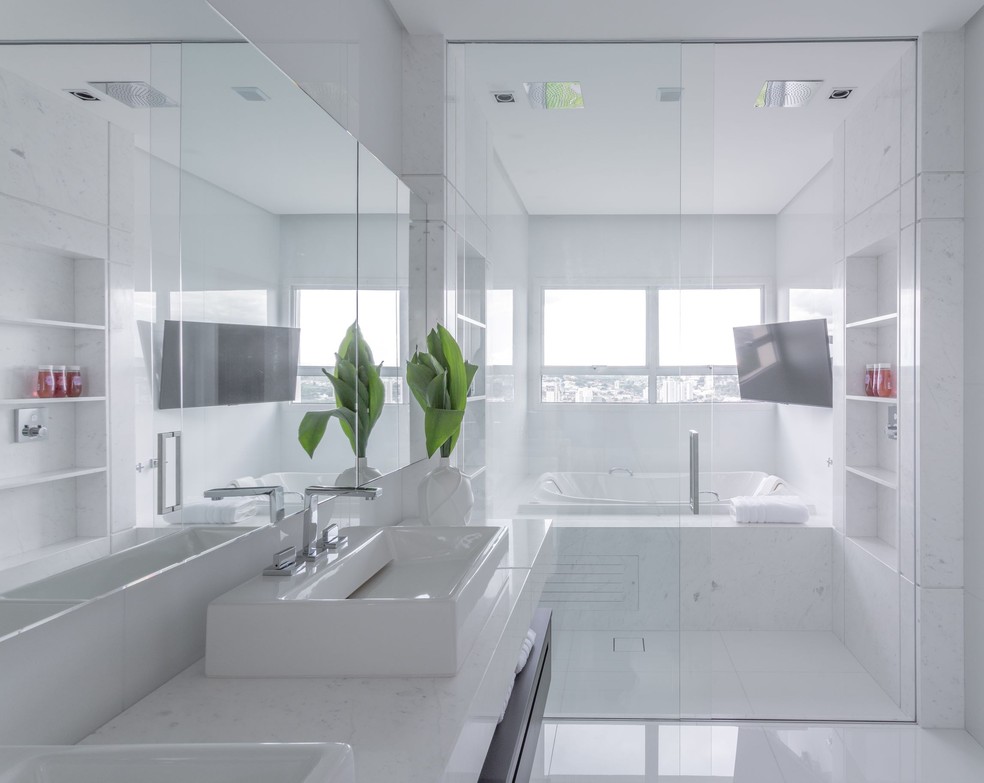 Além de revestir a base da banheira, o mármore branco forma estantes ao lado de cada um dos chuveiros de teto. Projeto do escritório Patrícia Penna Arquitetura & Design — Foto: Leandro Moraes