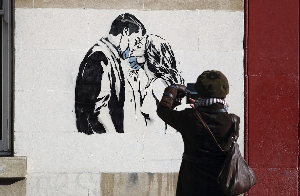 Mulher fotografa um novo mural surgido na Bank Street em Glasgow, na Escócia, que mostra um casal abaixando as máscaras para trocar um beijo. De autoria do artista de rua conhecido como Rebel Bear, a obra é inspirada em barreiras provocadas ela pandemia do coronavírus — Foto: Andrew Milligan / PA via AP