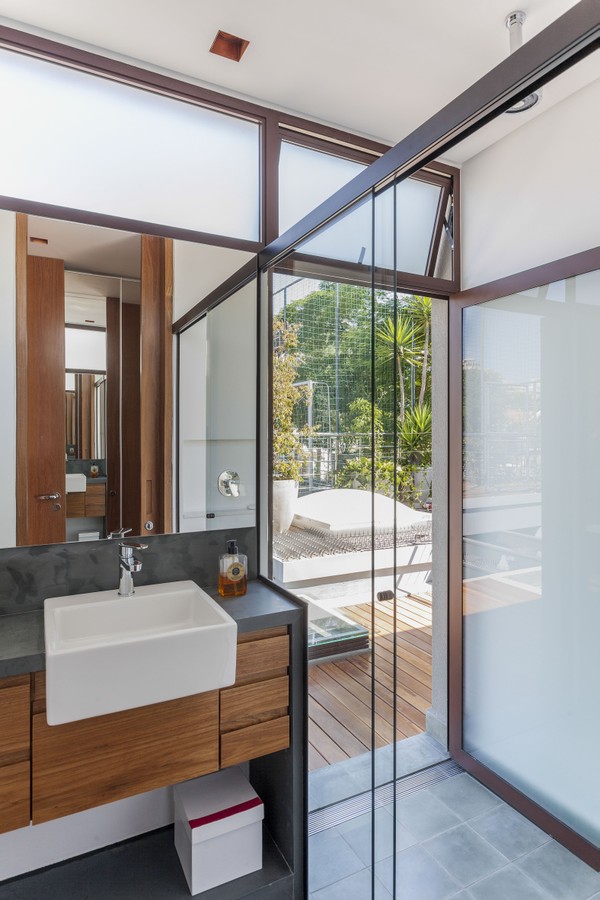 banheiro com box de vidro, bancada de cimento queimado com cuba de sobrepor branca, armário de madeira, espelho quadrado e piso cinza.