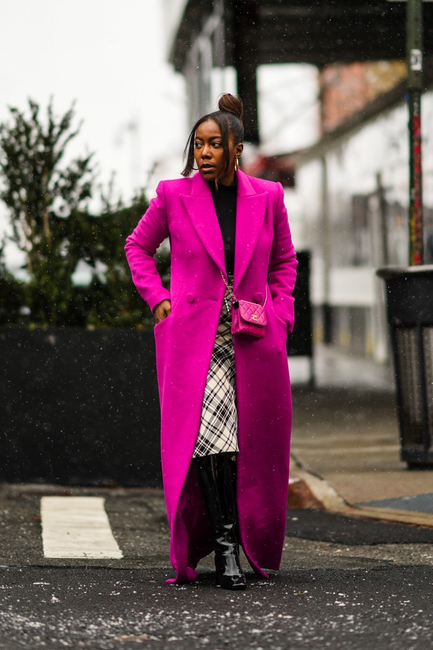 NEW YORK, NEW YORK - FEBRUARY 13: A guest wears gold large earrings, a black turtleneck pullover, a neon pink long coat, a pink shiny leather crossbody bag from Chanel, a white with black checkered print pattern midi skirt, black shiny varnished leather b (Foto: Getty Images)