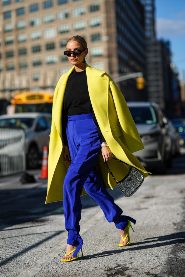 NEW YORK, NEW YORK - FEBRUARY 14: Leonie Hanne wears black sunglasses, a black and silver Prada pendant earring, a black high neck wool pullover, a yellow oversized long coat, high waist royal blue electric cargo large pants, a black leather and embroider (Foto: Getty Images)