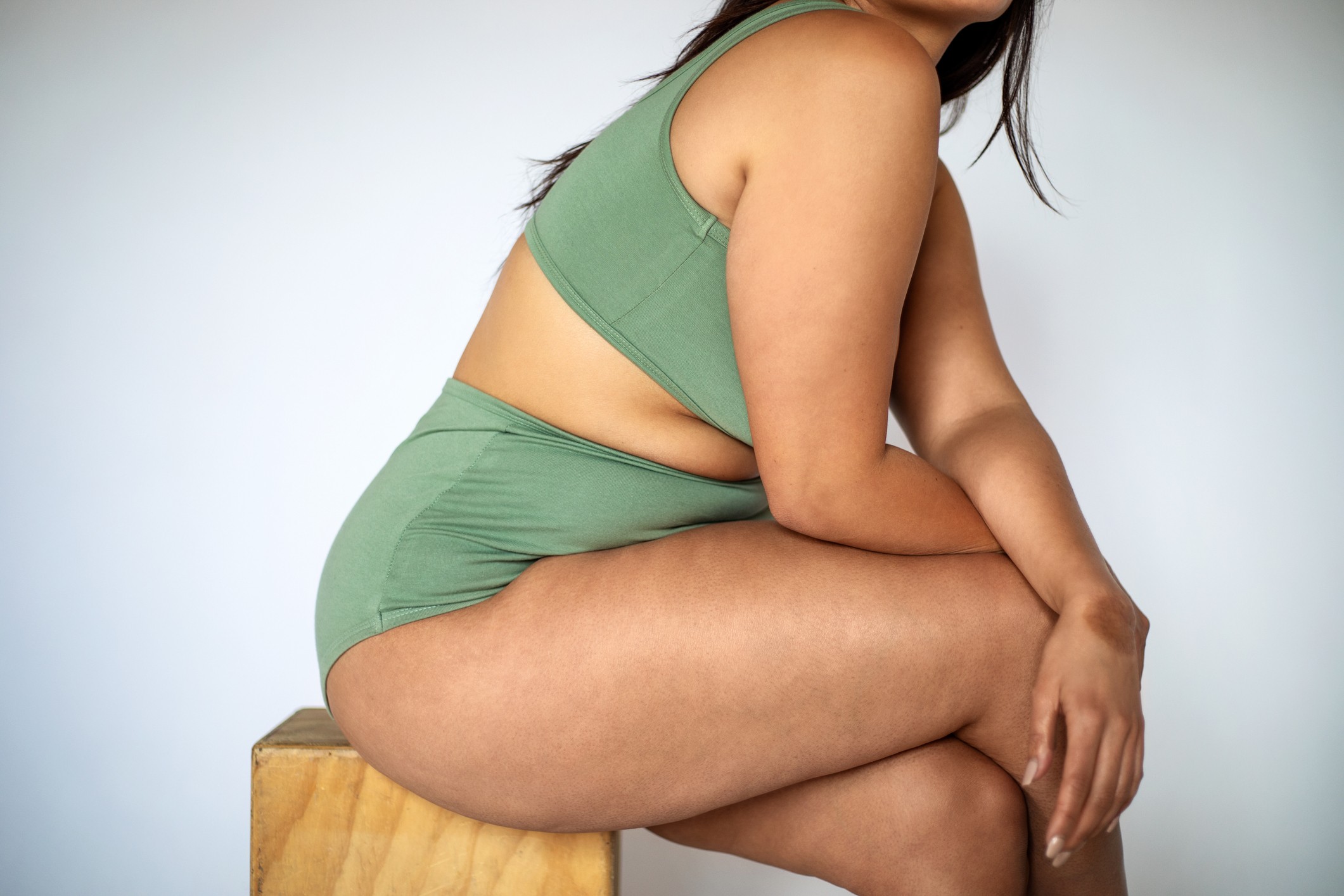 Plus size woman with fat legs sitting on stool. Oversized woman wearing lingerie with cellulite on thighs over white background. (Foto: Getty Images)