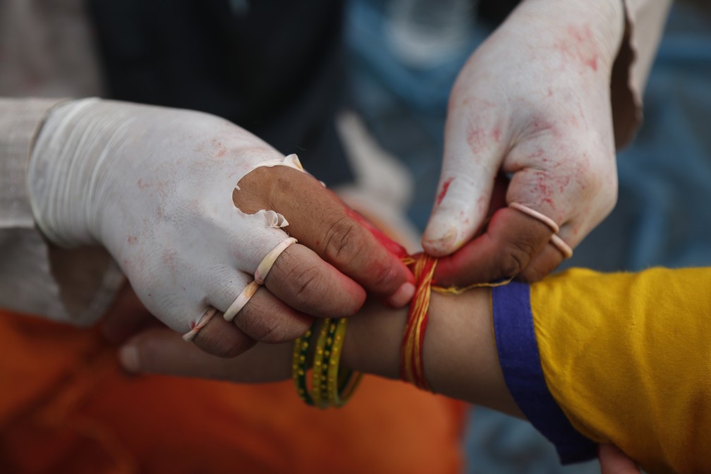 3 de agosto - Sacerdote hindu amarra o Janai, ou fio sagrado, no pulso de devotos mesmo depois que as luvas cirúrgicas rasgaram durante o festival Janai Purnima, em Kathmandu, Nepal. O sacerdote usava as luvas como precaução contra o coronavírus — Foto: Niranjan Shrestha/AP